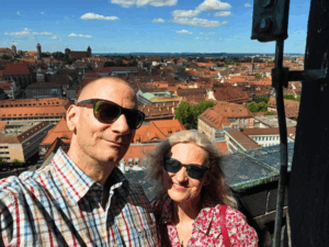 Ein Mann mit Glatz und eine Frau mit blonden Haaren und Sommerkleid, beide mit Sonnenbrille, lächeln vor dem Ausblick vom Turm der Lorenzkirche auf ganz Nürnberg.